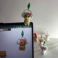 Toy figure of a bear with a green leaf on a white wall, with a computer screen displaying similar toy figure.