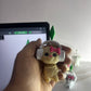 Hand holding a small plush toy with headphones and a bow tie in front of a computer screen.